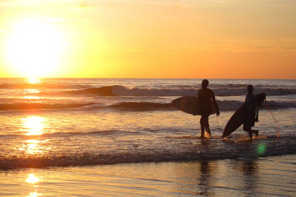 Dos surfistas caminan por la playa al atardecer en Nuquí, destino de surf en el Pacífico colombiano.