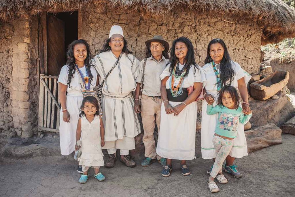 Familia indígena arhuaca con vestimenta típica, frente a una vivienda tradicional en la Sierra Nevada de Santa Marta.