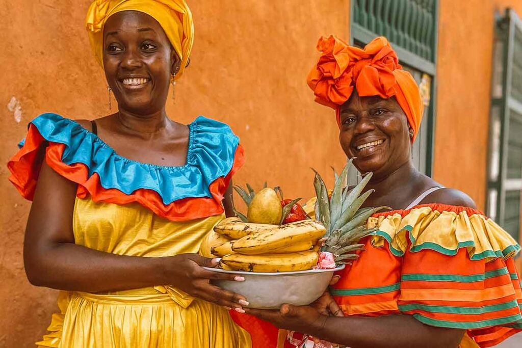 Palenqueras en Cartagena vestidas con trajes vibrantes, sosteniendo bandeja con frutas tropicales.